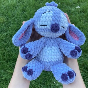 May include: A handmade blue plush toy with large ears, a purple nose, and dark purple accents on the feet. The toy is made of soft, textured yarn and has closed eyes, giving it a sleepy appearance. The background is green grass.