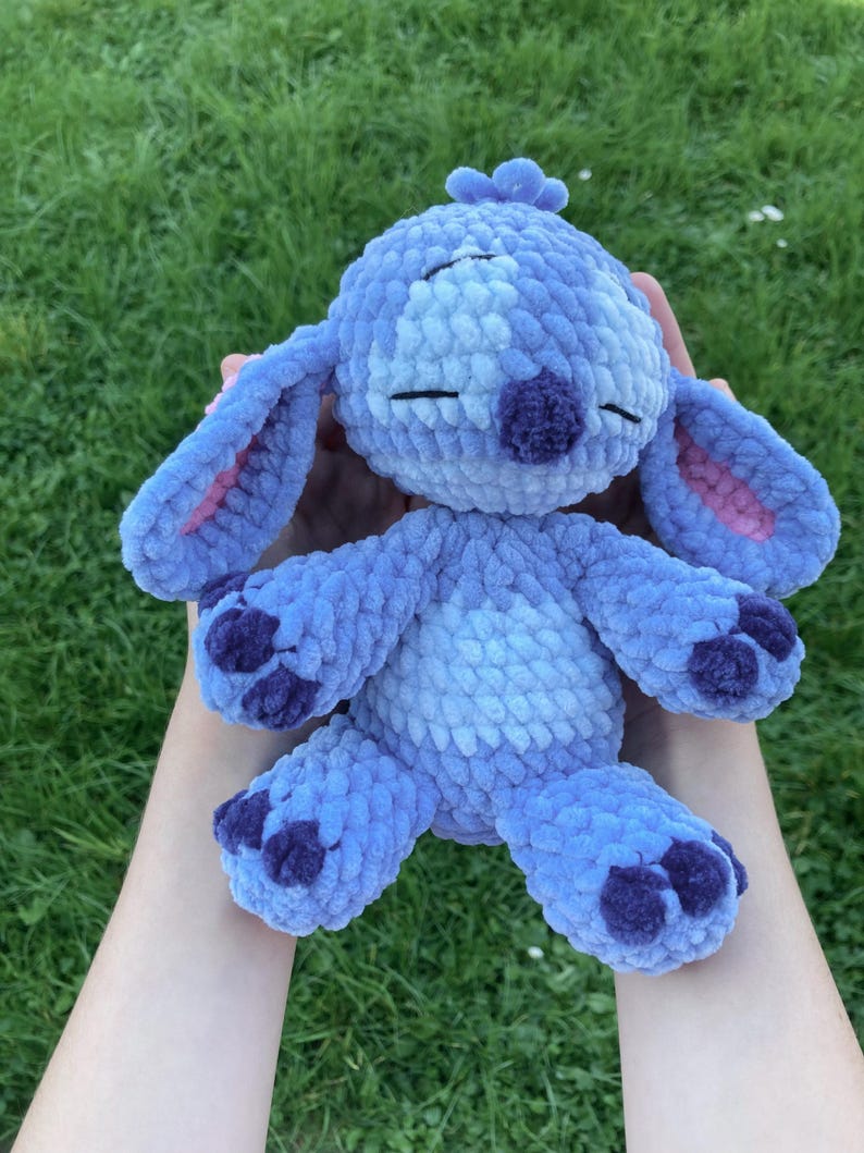 May include: A handmade blue plush toy with large ears, a purple nose, and dark purple accents on the feet. The toy is made of soft, textured yarn and has closed eyes, giving it a sleepy appearance. The background is green grass.