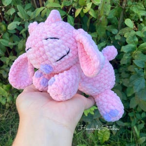 May include: A handmade pink crochet plush toy with a smiling face, large ears, and small purple accents on the feet and nose. The text "Heavenly Stitches" is visible in the bottom right corner. The plush is held in a hand against a background of green foliage.