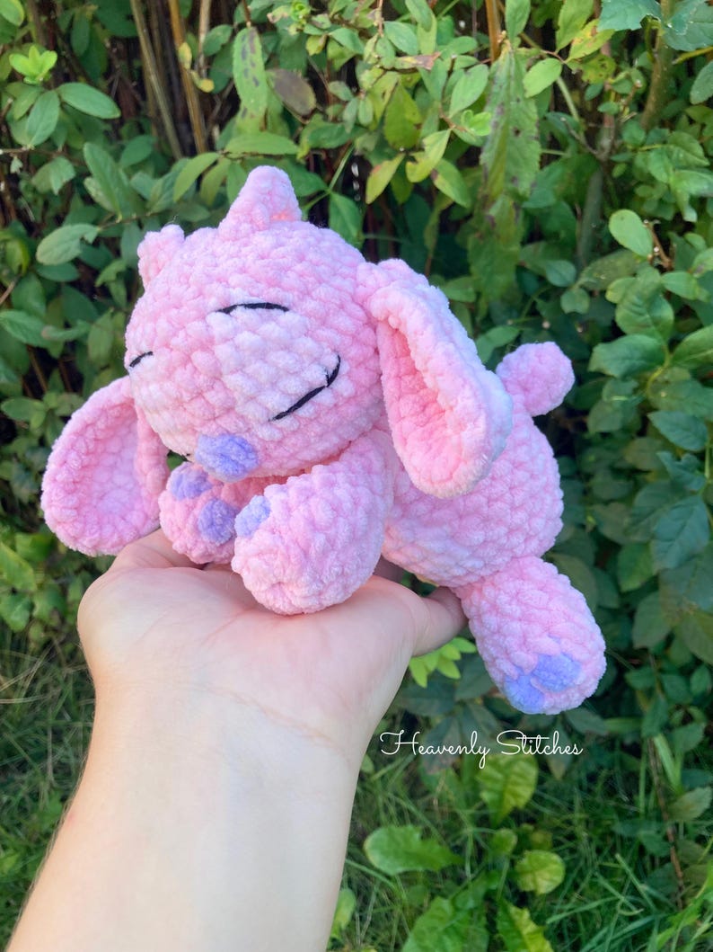 May include: A handmade pink crochet plush toy with a smiling face, large ears, and small purple accents on the feet and nose. The text "Heavenly Stitches" is visible in the bottom right corner. The plush is held in a hand against a background of green foliage.