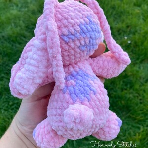 May include: A handmade pink and purple crochet bunny plushie. The plushie has long ears, a small tail, and purple accents on its head and back. The text "Heavenly Stitches" is written in cursive on the bottom right.