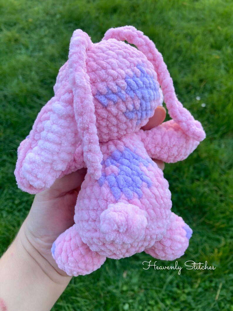 May include: A handmade pink and purple crochet bunny plushie. The plushie has long ears, a small tail, and purple accents on its head and back. The text "Heavenly Stitches" is written in cursive on the bottom right.