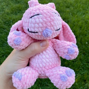 May include: A handmade pink crochet bunny with lavender accents on the paws and nose. The bunny has closed eyes and long ears. The text "Heavenly Stitches" is at the bottom of the image.