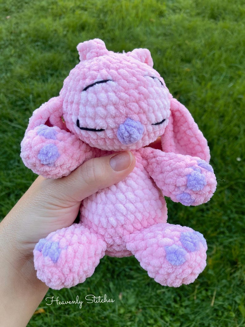 May include: A handmade pink crochet bunny with lavender accents on the paws and nose. The bunny has closed eyes and long ears. The text "Heavenly Stitches" is at the bottom of the image.