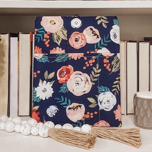 以下が含まれることがあります： ピンク、赤、白の花が咲いた花柄のネイビーブルーの布製ブックカバー。カバーには前面にポケットとボタン留めがあります。
