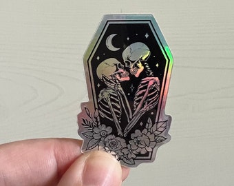 May include: A holographic sticker featuring a couple of skeletons embracing inside a coffin. The skeletons are kissing and surrounded by flowers. The sticker has a black background with a crescent moon and stars.