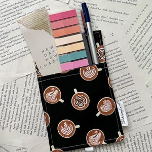 May include: A black fabric pouch with a zipper closure and a white zipper pull. The pouch is decorated with a repeating pattern of brown coffee cups with white latte art. The pouch is sitting on top of a stack of papers with colorful sticky notes and a silver pen.