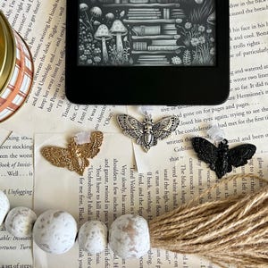 May include: Three silver and gold butterfly book markers with intricate details. The markers are on a background of a book with text and a brown woven surface.