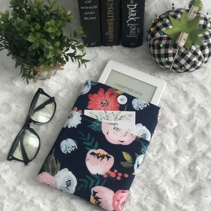 以下が含まれることがあります： ピンク、赤、白の花柄が施されたネイビーブルーの電子書籍リーダーカバー。カバーの前面には、名刺やブックマークを入れるためのポケットがあります。カードには「ringCo」の文字が見えます。