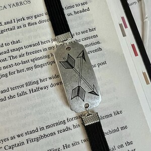 May include: A black bookmark with a silver charm featuring two crossed arrows. The charm has a rustic, aged look. The bookmark is made of a soft, flexible material.