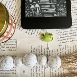May include: A close-up of a green frog bookmark with a white background. The bookmark is attached to a book with a black e-reader on top of it. The book has a white background with brown text. There are white and brown beads on the book.