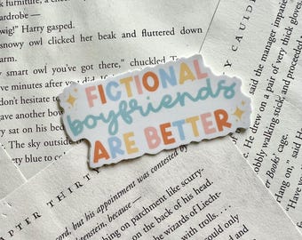 May include: A white sticker with the text "FICTIONAL BOYFRIENDS ARE BETTER" in colorful letters. The sticker has a cloud-like shape and is decorated with small starbursts. The sticker is placed on top of open book pages.