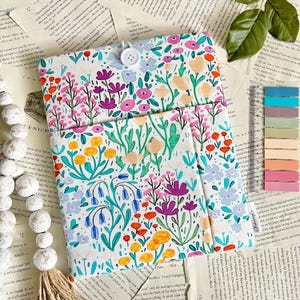 May include: A floral print book cover with a button closure. The cover features a white background with colorful flowers in shades of pink, purple, orange, and blue. The book cover is open, revealing pages of a book.