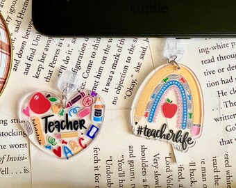 May include: Two acrylic teacher appreciation keychains. One is a heart shape with the word "Teacher" and school supplies inside. The other is a rainbow with a red apple and the hashtag "#teacherlife".