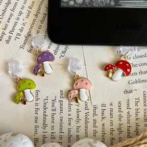 May include: A set of four mushroom earrings in various colors: green, purple, pink, and red. Each earring features a gold-colored stem and a white base. The earrings are designed with a whimsical, nature-inspired theme.