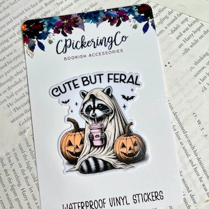 May include: Waterproof vinyl sticker featuring a cute raccoon wearing a ghost costume and holding a pink drink. The sticker has the text "Cute But Feral" and is surrounded by bats and pumpkins.