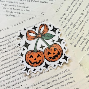 May include: A Halloween-themed sticker featuring two orange jack-o'-lanterns with smiling faces, connected by a green stem and a decorative orange bow. The sticker is surrounded by black starbursts.