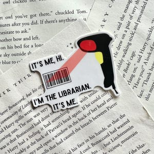 以下が含まれることがあります： 「IT'S ME, HI. I'M THE LIBRARIAN. IT'S ME.」の文字が入った、バーコードスキャナーの形をした白いステッカー。スキャナーは黒色で、赤と黄色のアクセントと赤いビームが付いています。ステッカーは本のページを背景にしています。