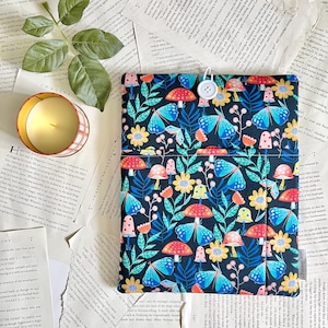 May include: A dark blue fabric book sleeve with a colorful pattern of butterflies, mushrooms, and flowers. The sleeve has a white button closure and a pocket. A lit candle and book pages are in the background.