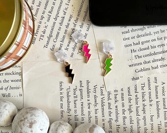 May include: Three gold lightning bolt earrings with black, pink, and green enamel. The earrings are on a white surface with a book and white beads.