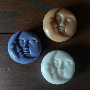 May include: Three round soaps featuring a crescent moon and face design. The soaps are in shades of brown, white, and purple.