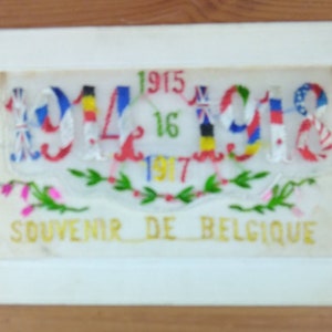 Puede incluir: Un recuerdo enmarcado de Bélgica, con texto y números bordados. Los números 1914-1918 se muestran con banderas coloridas, y el texto "SOUVENIR DE BELGIQUE" es visible. El marco es blanco.