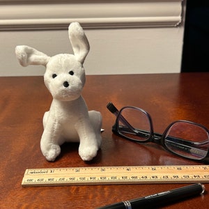 May include: A white plush toy dog with black eyes and a black nose. The dog is sitting on a wooden table with a ruler, a pen, and a pair of glasses.