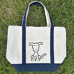 May include: Cream-colored canvas tote bag with navy blue accents. The bag features a black line drawing of a dog on the front, a navy blue base, and navy blue handles. The bag has a zipper closure and a small front pocket.