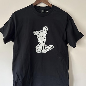 May include: Black t-shirt featuring a white cartoon dog design. The dog is sitting with its tongue out. The shirt is hanging on a wooden hanger against a white wall.