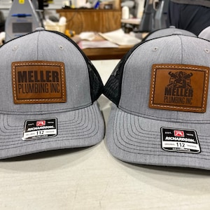 May include: Two gray and black trucker hats with brown leather patches. The patches have the text "Meller Plumbing Inc." and a graphic of a person lifting weights. The hats have a black mesh back and a Richardson label.