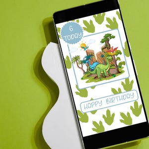 May include: A smartphone displays a birthday card design with a dinosaur theme. The card features a green and blue dinosaur illustration, the text "Happy Birthday," and the number "6 Today." The phone rests on a white, abstract-shaped stand against a green background.