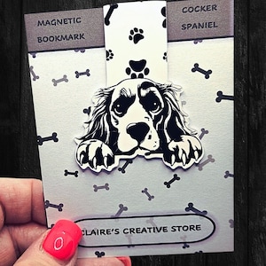 May include: A white magnetic bookmark featuring a black and white illustration of a Cocker Spaniel dog peeking over the top. The bookmark has a white background with black paw prints and bones. The text "MAGNETIC BOOKMARK" is printed on the left side of the bookmark and "COCKER SPANIEL" is printed on the right side. The text "CLAIRE'S CREATIVE STORE" is printed on the bottom of the bookmark.