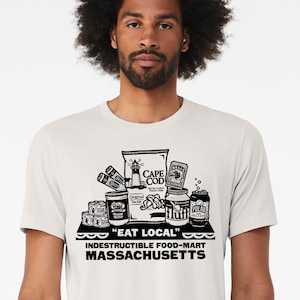 May include: A light-colored t-shirt featuring a black and white graphic of various food items, including a bag of chips and cans, with the text "EAT LOCAL" and "INDESTUCTIBLE FOOD-MART MASSACHUSETTS".