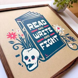 May include: A framed art print with a brown wooden frame. The print features a teal book with the words "READ AND WRITE AND FIGHT" in white and red. A white skull, pencils, and floral elements complete the design.