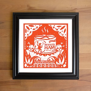 May include: A framed print featuring a vibrant orange and white design. The artwork showcases a can of "Deviled Ham" with a devilish character, skulls, and decorative elements. The print is set against a wooden background.