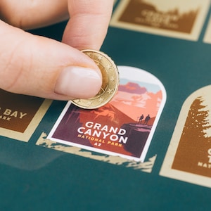 Pode incluir: Uma m&atilde;o usando uma moeda para raspar um adesivo do Parque Nacional do Grand Canyon em um fundo verde-azulado. O adesivo apresenta uma ilustra&ccedil;&atilde;o da paisagem com o texto "GRAND CANYON NATIONAL PARK AZ". Outros adesivos s&atilde;o vis&iacute;veis.