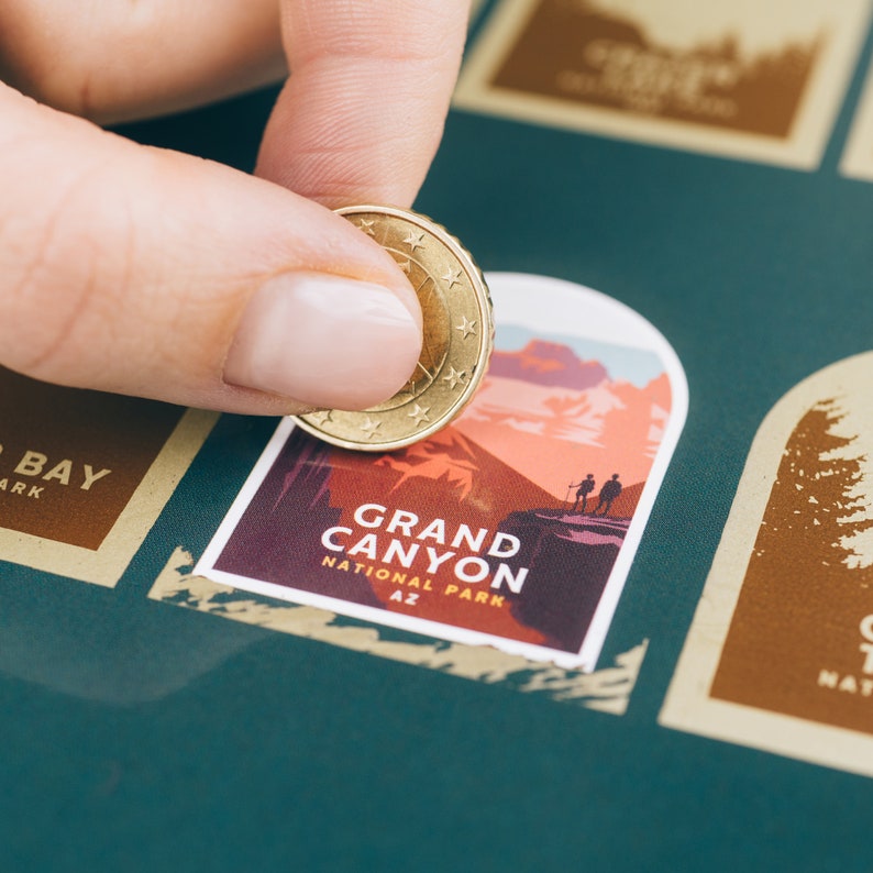 Pode incluir: Uma m&atilde;o usando uma moeda para raspar um adesivo do Parque Nacional do Grand Canyon em um fundo verde-azulado. O adesivo apresenta uma ilustra&ccedil;&atilde;o da paisagem com o texto "GRAND CANYON NATIONAL PARK AZ". Outros adesivos s&atilde;o vis&iacute;veis.