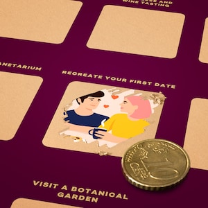 Puede incluir: Un tablero de actividades raspable morado y dorado con el texto "RECREATE YOUR FIRST DATE" y una ilustraci&oacute;n de una pareja. Una moneda est&aacute; en primer plano. El tablero tiene otros cuadrados de actividad.
