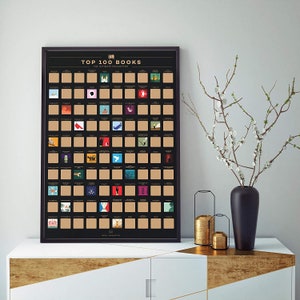 Könnte beinhalten: Ein gerahmtes schwarzes Poster mit dem Titel "TOP 100 BOOKS" mit Rubbelfeldern. Das Poster zeigt ein Raster von Buchcovern, von denen einige freigelegt sind. Das Poster wird auf einem weißen Schrank mit goldenen Akzenten neben einer Vase mit weißen Blumen ausgestellt.