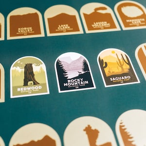 Pode incluir: Uma cole&ccedil;&atilde;o de autocolantes arqueados com ilustra&ccedil;&otilde;es de parques nacionais. Os autocolantes apresentam paisagens como Redwood, Rocky Mountain e Saguaro National Parks. Cada autocolante inclui o nome do parque e a abrevia&ccedil;&atilde;o do estado, num fundo verde-azulado.