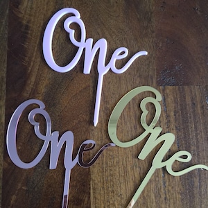Può includere: Tre topper per torta che compongono la parola "Uno" in stili diversi. I topper sono in acrilico e sono rosa, oro rosa e oro.