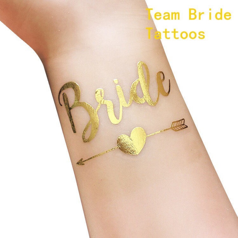 May include: Gold temporary tattoo with the word "Bride" in a cursive font and an arrow with a heart. The text "Team Bride Tattoos" is at the top of the image.