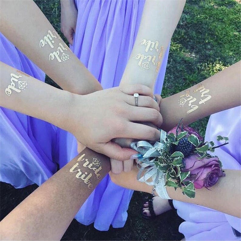 May include: Four people wearing temporary tattoos that say "Bride Tribe" in gold lettering with a diamond design. The tattoos are on their arms and wrists. The people are wearing purple dresses and are holding hands.