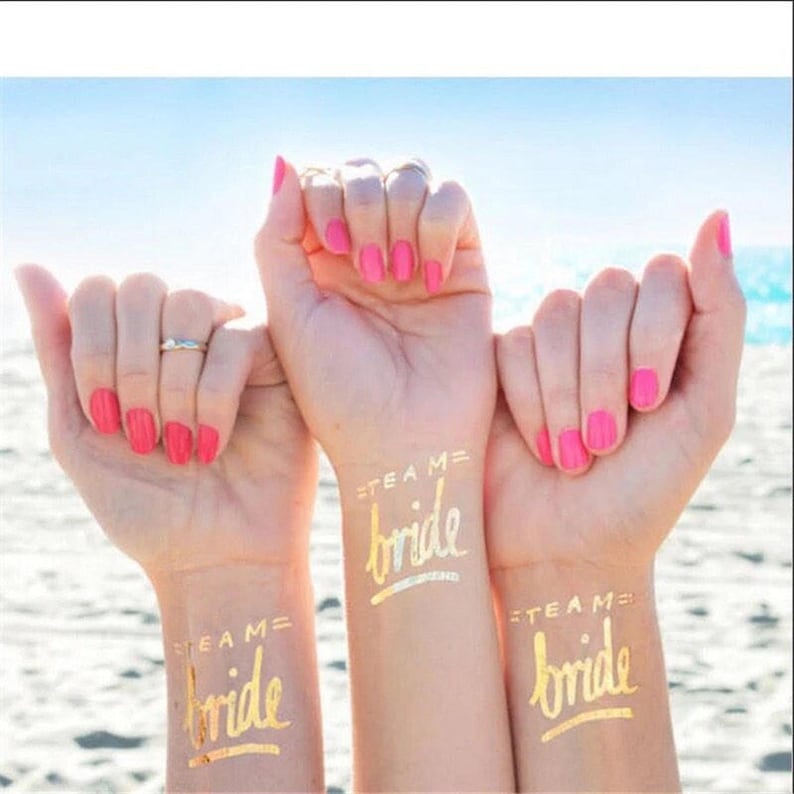 May include: Three people's arms with gold temporary tattoos that say "Team Bride" on their wrists. The tattoos are in a cursive font.