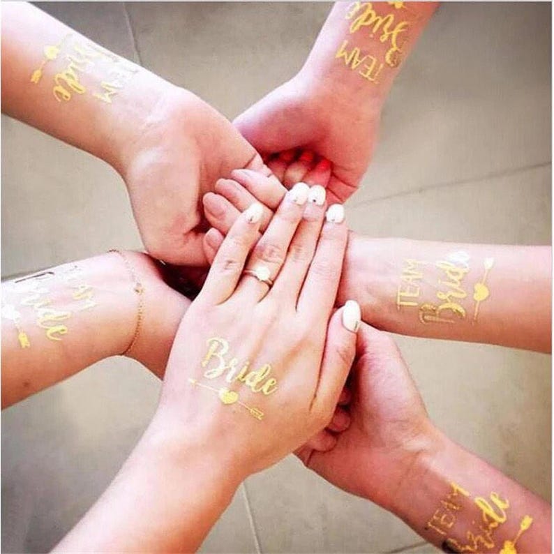 May include: Gold temporary tattoos with the words "Team Bride" and "Bride" on them. The tattoos are on the wrists of several people.