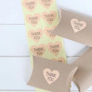 Puede incluir: Un rollo de pegatinas amarillas con forma de corazón color melocotón, cada una con las palabras "THANK YOU" en negro. También se ven varias cajas de almohada marrones pequeñas con pegatinas de corazón a juego.