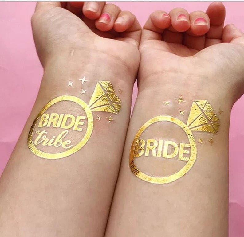 May include: Two gold temporary tattoos on a person's wrists. The tattoos are in the shape of a ring with a diamond on top. One tattoo says "bride tribe" and the other says "bride".