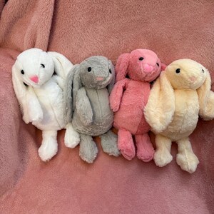 Op de afbeelding: Vier pluche konijnen speelgoed in verschillende kleuren: wit, grijs, roze en geel. De konijnen hebben lange, slappe oren en zitten op een roze, pluche oppervlak.