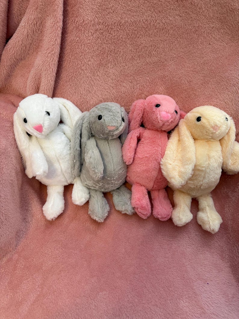 Op de afbeelding: Vier pluche konijnen speelgoed in verschillende kleuren: wit, grijs, roze en geel. De konijnen hebben lange, slappe oren en zitten op een roze, pluche oppervlak.