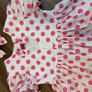 Op de afbeelding: Een witte jurk met roze stippen en een knoopsluiting op de rug. De jurk heeft korte mouwen en een volle rok. Een roze strik is inbegrepen.